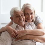 White-haired couple hugging and smiling on a couch in a well-lit room